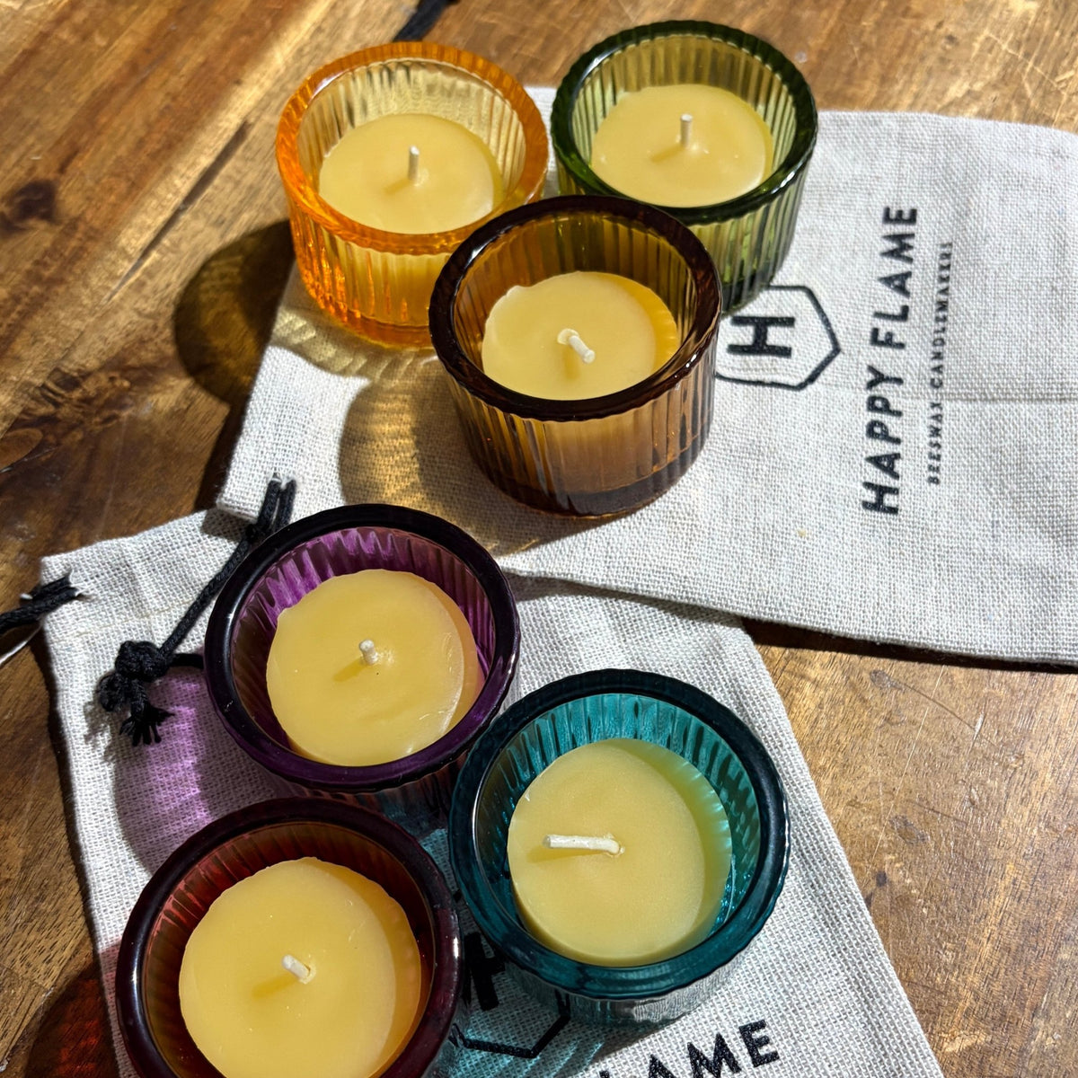 Six small beeswax candles in colorful glass holders in a 'Happy Flame' linen bag