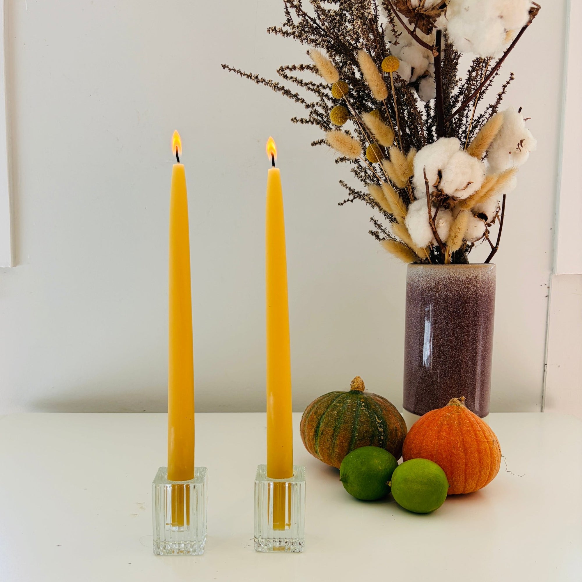 Two yellow candles in glass holders with pumpkins and a vase of dried flowers on a white surface.