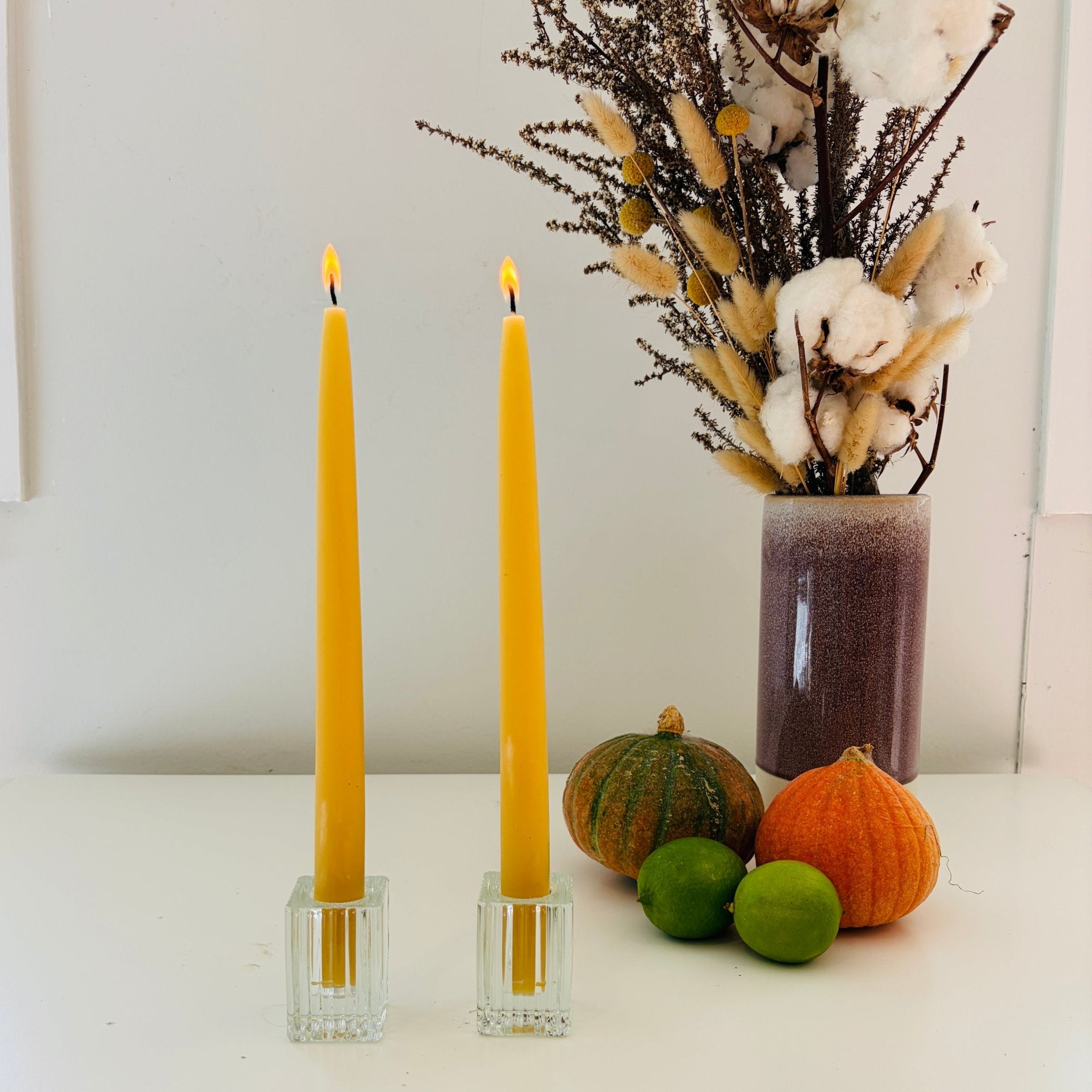 Two yellow candles in glass holders with pumpkins and a vase of dried flowers on a white surface.