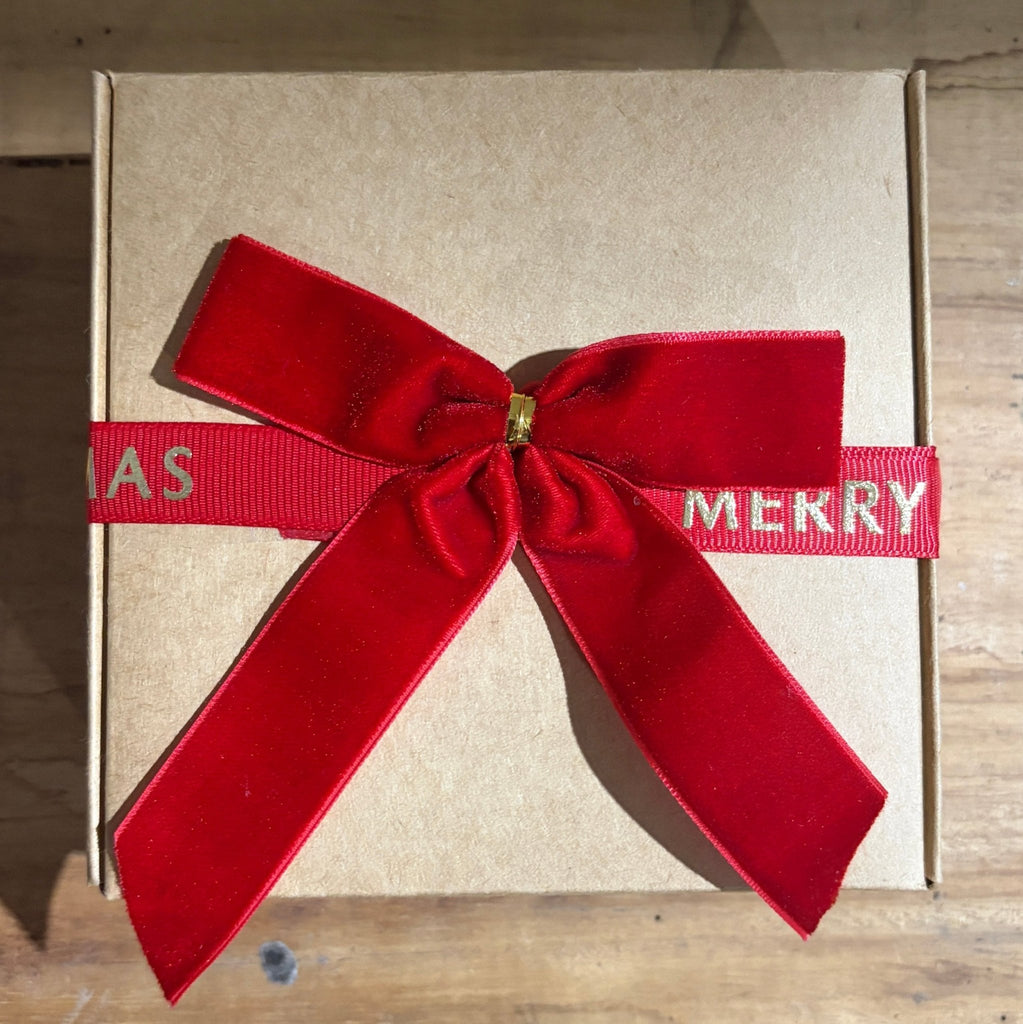 A kraft paper gift box tied with a red velvet ribbon bow and a red ribbon banner that reads 'MERRY CHRISTMAS' in white lettering, placed on a wooden surface.