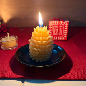 A yellow pinecone-shaped candle on a small black plate, lit with a flame, set on a red cloth with a small red patterned box in the background.