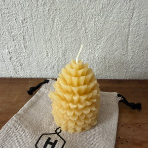 A beeswax candle in a pinecone shape sits on a beige drawstring bag with a black logo, placed on a wooden surface against a textured white wall.