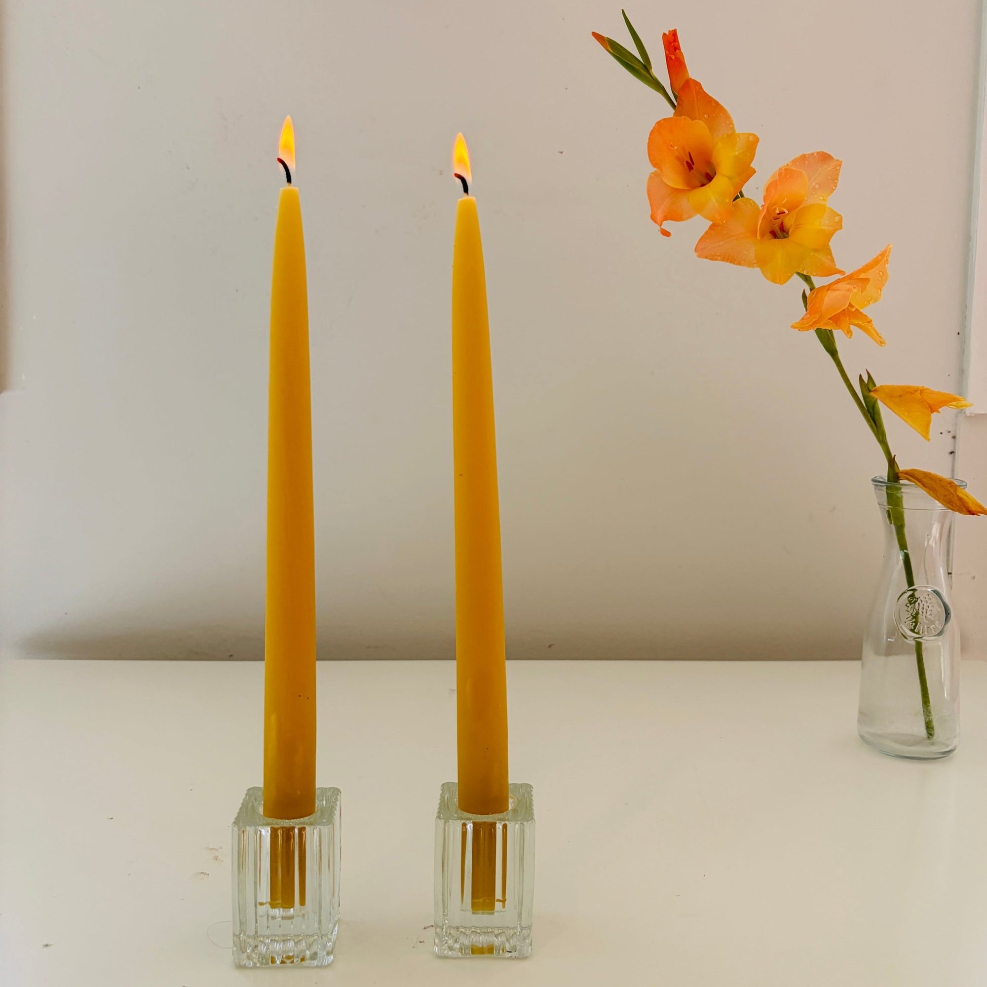 Two yellow candles in clear glass holders on a white surface with a vase of flowers in the background.