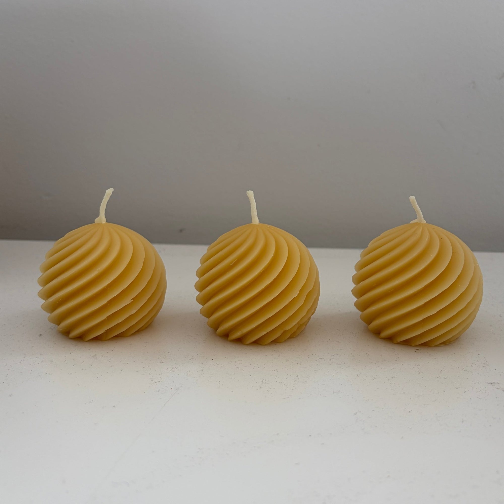 Three round beeswax candles with a ribbed spiral texture, arranged in a row on a white surface against a gray background.